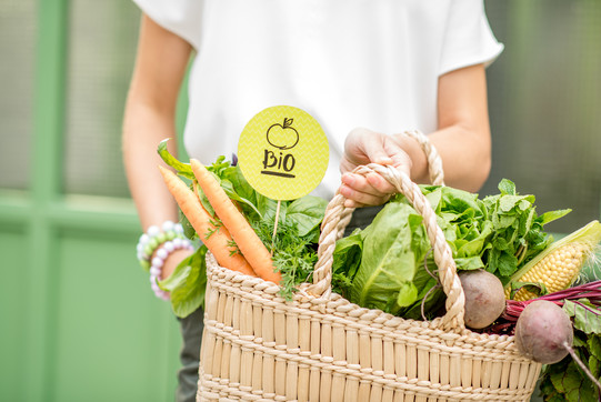 Eine Person mit weißem T-Shirt trägt einen mit Obst und Gemüse gefüllten Einkaufskorb.