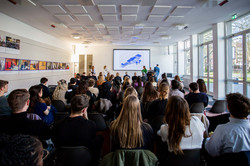 The picture shows a seminar room with rows of chairs in which the audience is sitting. A screen with a presentation picture can be seen on the other side of the seminar room.