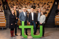 Vier Männer und zwei Frauen stehen vor den zwei grünen Buchstaben t und u. Im Hintergrund ist ein Hörsaal zu sehen, in dem mehrere Personen sitzen.