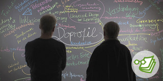 Man sieht zwei Männer von hinten, die vor einer Tafel stehen, auf der mehrere Begriffe stehen. In der Mitte steht das Wort Doprofil.