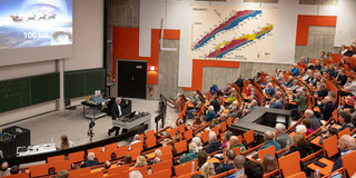 In einem gut besuchten Hörsaal mit orangenen Sitzen hält Prof. Metin Tolan einen Vortrag. Auf der Leinwand ist eine Präsentation zu sehen.
