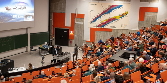 In einem gut besuchten Hörsaal mit orangenen Sitzen hält Prof. Metin Tolan einen Vortrag. Auf der Leinwand ist eine Präsentation zu sehen.