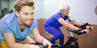 Eine ältere Frau und ein junger Mann sitzen nebeneinander auf Spinning-Fahrrädern.