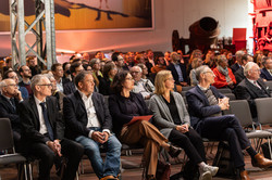 Ein Publikum in formeller Kleidung sitzt in einem Saal und schaut nach rechts.