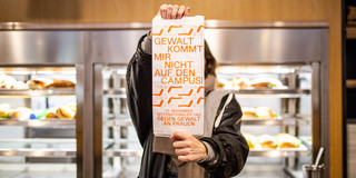 A young woman stands in front of a canteen shelf filled with sandwiches and holds a sandwich bag with the words "Violence doesn't come on campus" in front of her face.