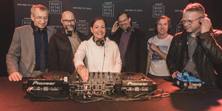 Sechs Personen stehen an einem DJ Pult. Sie tragen Kopfhörer.