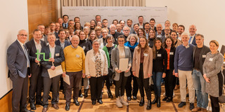 Stipendienfeier Eine Gruppe von Menschen steht zusammen. Zwei Personen auf der linken Seite der Gruppe halten die Buchstaben t und u in die Höhe.