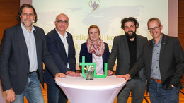 Das Bild zeigt Prof. Andreas Löschel, Prof. Manfred Bayer, Prof. Petra Wiederkehr, Prof. Aladin El-Mafaalani, Markus Neuhaus (v.l.n.r.) beim Gambrinus-Forum. In der Mitte vor ihnen steht ein weißer Stehtisch mit dem grünen TU-Logo. Im Hintergrund ist eine Präsentation zu sehen.