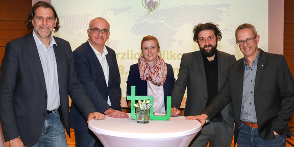 Das Bild zeigt Prof. Andreas Löschel, Prof. Manfred Bayer, Prof. Petra Wiederkehr, Prof. Aladin El-Mafaalani, Markus Neuhaus (v.l.n.r.) beim Gambrinus-Forum. In der Mitte vor ihnen steht ein weißer Stehtisch mit dem grünen TU-Logo. Im Hintergrund ist eine Präsentation zu sehen.