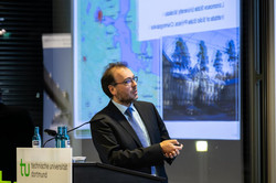 A man in a suit with a light blue shirt and tie gives a presentation.