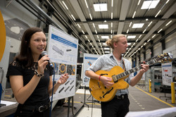 Ein Mann und eine Frau stehen in der Versuchshalle. Der Mann spielt Gitarre, die Frau singt. 