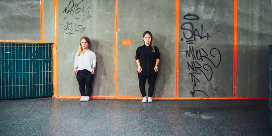 Zwei kleinwüchsige Frauen stehen vor einer Betonwand und tragen schlichte Kleidung der Marke "Auf Augenhöhe".