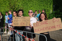 Motivationsposter Am Rande der Laufstrecke stehen zwei Personen, die Schilder für die Läufer*innen hochhalten. Auf Schild 1 steht: "Running away from your ex?" Auf Schild 2 steht: "Leonie, du Zuckerschnute"