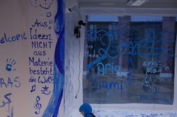 Ein Kind malt mit blauer Farbe und einem Pinsel auf eine Wand, die bereits bemalt wurde