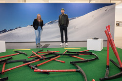 Zwei Personen stehen vor einer Fotowand mit einem verschneiten Bergpanorama. Auf dem Boden sind rote und schwarze Metallstangen auf grünem Untergrund verteilt, die wie demontierte Teile von Wintersportgeräten wirken.