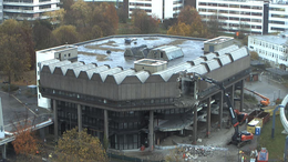 Das Bild zeigt die alte Bibliothek der TU Dortmund vom Mathetower aus. Im Hintergrund sind die Gebäude der FH Dortmund zu sehen. Rechts neben der ehemaligen Zentralbibliothek befinden sich Bagger, die die Fassade des Gebäudes abtragen.