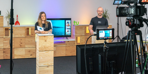 A woman and a man stand in front of two boxes and moderate an event. A camera is filming them.