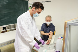 A man in a white doctor's coat draws blood from another man.