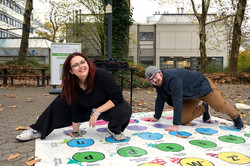 Das Bild zeigt zwei erwachsene Personen auf dem Campus, die auf einer Art "Twister-Plane" hocken und ihren linken Fuß sowie ihre rechte Hand auf kreisrunde Felder gelegt haben.
