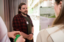 In front of a young man with long brown hair and a beard who is explaining something, a man holding two green stress balls stands to the left and a young woman with glasses stands to the right.