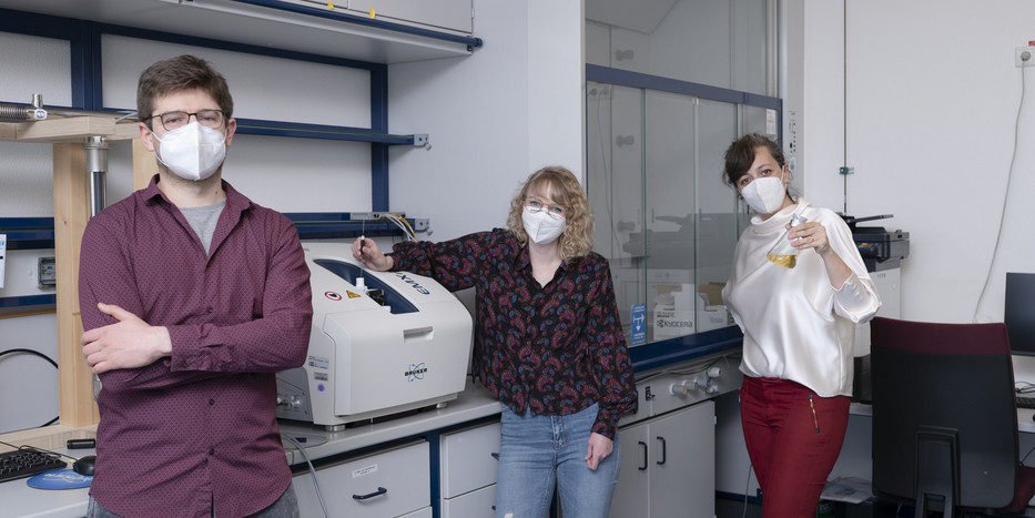 Drei Personen stehen in einem Chemie-Labor. Die Frau rechts hält einen Erlenmeyerkolben mit gelber Flüssigkeit in der Hand.