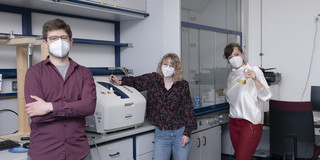 Drei Personen stehen in einem Chemie-Labor. Die Frau rechts hält einen Erlenmeyerkolben mit gelber Flüssigkeit in der Hand.