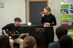 Ein junger Mann sitzt links im Bild und spielt Gitarre. Rechts neben ihm steht eine junge Frau und singt in ein Mikrofon.