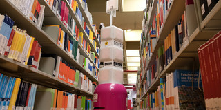 A service robot stands between two bookshelves in the university library.