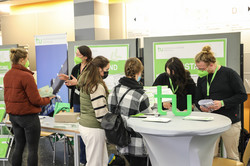 Several women and men are standing in front of a TU Dortmund booth looking at material.