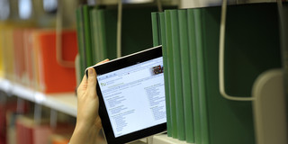Das Bild zeigt ein Tablet mit leuchtendem Bildschirm, das in eine Reihe von Büchern in der Bibliothek gehalten wird.
