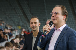 Im Vordergrund ein Mann der ein Mikrofon in der Hand hält, dahinter ein weiterer Mann und die volle Tribüne des BVB-Stadions.