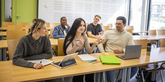 Eine Gruppe fünf Studierender sitz auf zwei Tischreihen aufgeteilt in einem Seminarraum. Auf der Tischreihe im Vordergrund liegen Schreibblöcke, Hefter und Laptops.