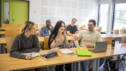 Eine Gruppe fünf Studierender sitz auf zwei Tischreihen aufgeteilt in einem Seminarraum. Auf der Tischreihe im Vordergrund liegen Schreibblöcke, Hefter und Laptops.