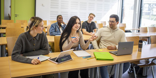 Eine Gruppe fünf Studierender sitz auf zwei Tischreihen aufgeteilt in einem Seminarraum. Auf der Tischreihe im Vordergrund liegen Schreibblöcke, Hefter und Laptops.