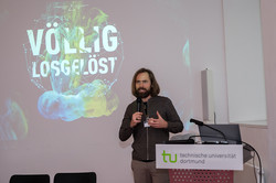 Das Bild zeigt einen Mann mit Bart, der schräg hinter einem Pult steht. Im Hintergrund ist die Präsentation "Völlig losgelöst" zu sehen.