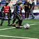 Foto: Drei Roboter in roten und blauen Trikots spielen Fußball. Der Roboter im blauen Trikot setzt zum Schuss an.
