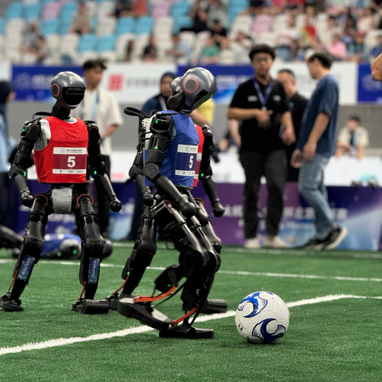 Foto: Drei Roboter in roten und blauen Trikots spielen Fußball. Der Roboter im blauen Trikot setzt zum Schuss an.