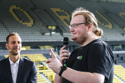 Ein Vertreter des AstA, der ein Mikrofon in der Hand hält. Neben ihm steht ein Mann im Anzug. Die beiden stehen auf einer Stadiontribüne des BVB-Stadions.