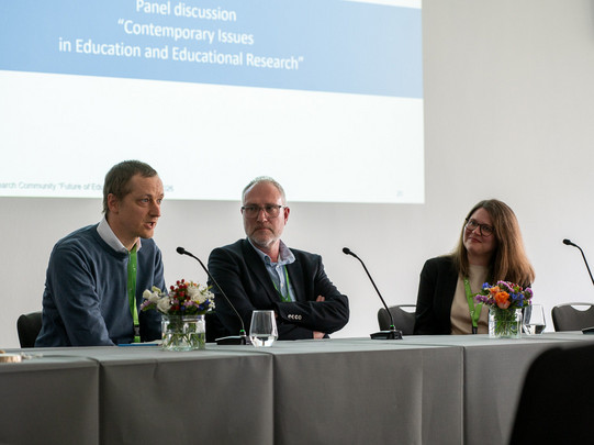 Foto: Drei Personen sitzen vorne im Tagungsraum am Konferenztisch und sprechen in Mikrofone