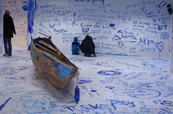 Ein bemaltes Holzboot steht in einem bemalten Raum. Menschen malen an die Wand