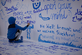 Ein Kind malt mit einem Pinsel und blauer Farbe an eine weiße Wand
