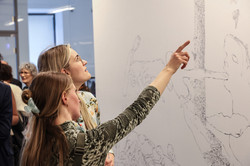 Im Fokus des Bildes stehen zwei Besucherinnen, die sich ein Ausstellungswerk angucken. Eine der Besucherin zeigt mit ausgestrecktem Arm auf dieses.