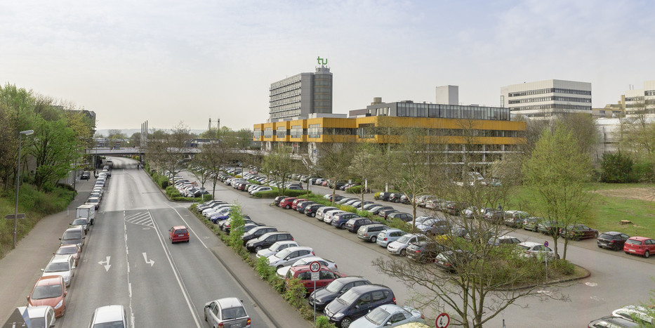 Ein Parkplatz voller Autos neben einer Straße, im Hintergrund ist das Mensagebäude und der Mathetower sowie weitere Gebäude der TU Dortmund zu sehen.
