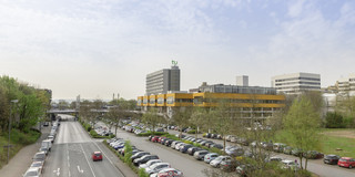 Ein Parkplatz voller Autos neben einer Straße, im Hintergrund ist das Mensagebäude und der Mathetower sowie weitere Gebäude der TU Dortmund zu sehen.