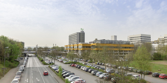 Parkplatz Ein Parkplatz voller Autos neben einer Straße, im Hintergrund ist das Mensagebäude und der Mathetower sowie weitere Gebäude der TU Dortmund zu sehen.