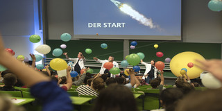 DLR Raumfahrtshow Schüler lassen im Hörsaal bunte Luftballons steigen, auf der Leinwand im Hintergrund ist der Start einer Rakete zu sehen