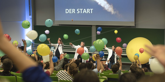 Schüler lassen im Hörsaal bunte Luftballons steigen, auf der Leinwand im Hintergrund ist der Start einer Rakete zu sehen