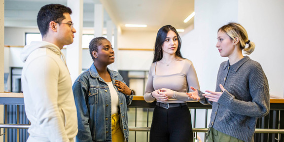 Das Bild zeigt vier Studierende, die in einem Halbkreis zueinander stehen. Drei Personen der Studierenden richten alle den Blick auf eine sprechende Person.