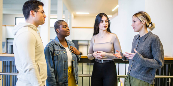 Das Bild zeigt vier Studierende, die in einem Halbkreis zueinander stehen. Drei Personen der Studierenden richten alle den Blick auf eine sprechende Person.