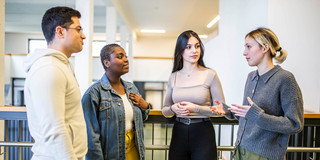 Das Bild zeigt vier Studierende, die in einem Halbkreis zueinander stehen. Drei Personen der Studierenden richten alle den Blick auf eine sprechende Person.